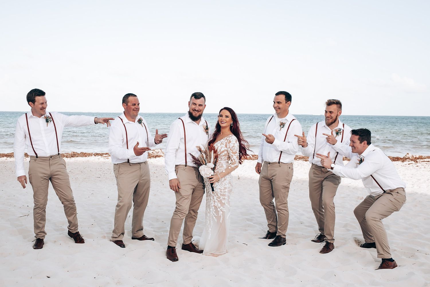 Wedding party performs synchronized dance moves on a sunny beach in matching khaki and white attire.