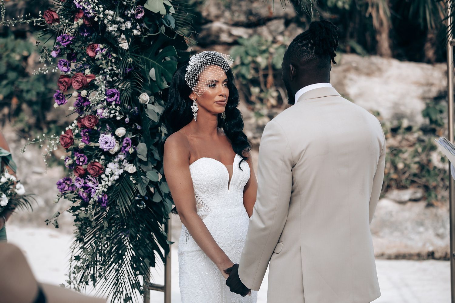 A romantic outdoor wedding ceremony with tropical foliage and purple floral decorations.