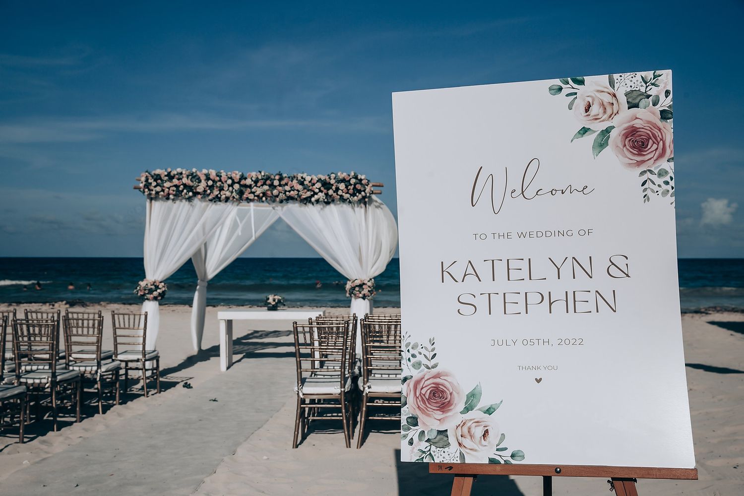 A beach wedding setup with white draped arch and floral decor next to a welcome sign with dusty rose accents.
