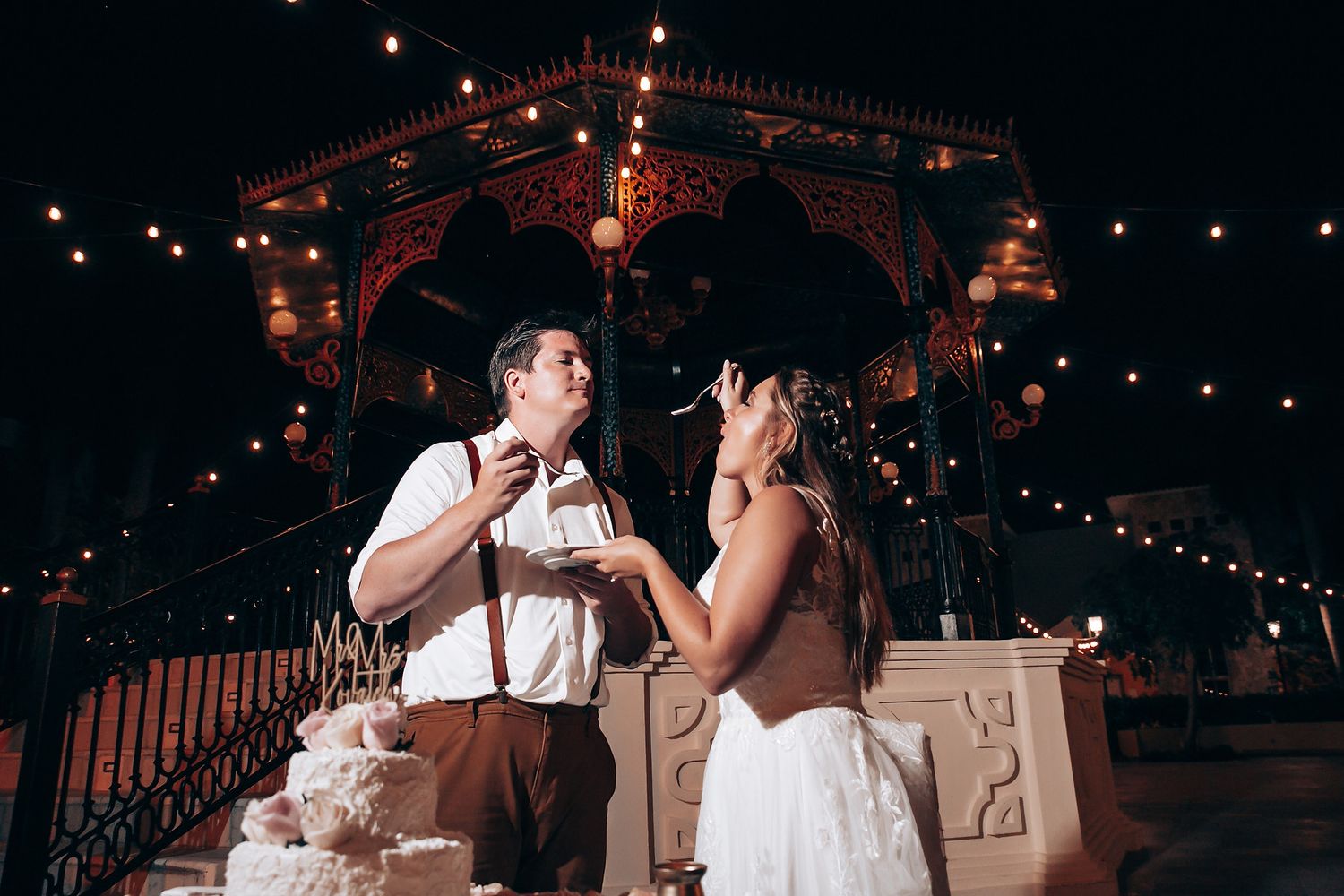 Wedding couple sharing a moment while cutting their cake under a decorated gazebo with string lights.