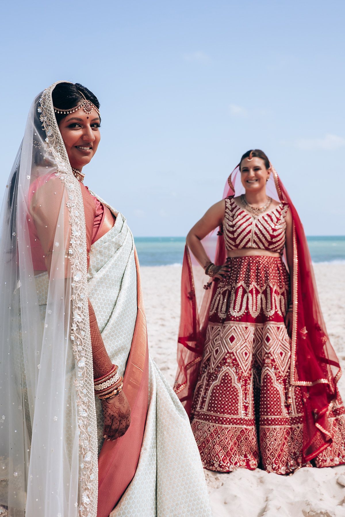 Two people wearing traditional Indian wedding attire pose on a beautiful white sandy beach.