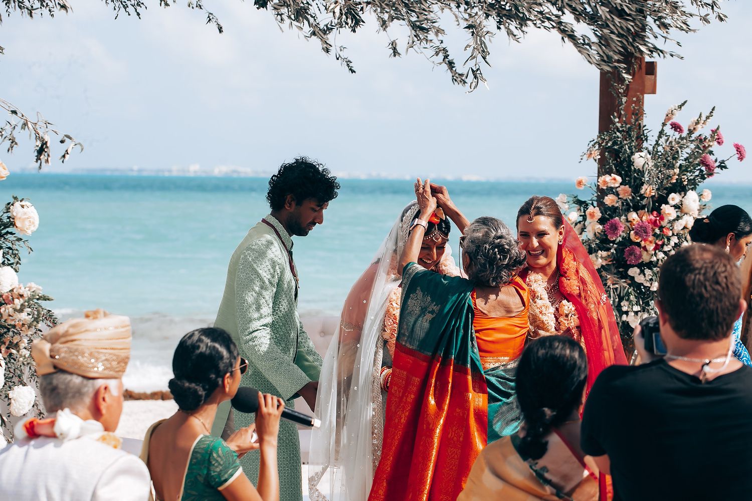 Beachside wedding ceremony with guests gathered under trees near turquoise waters.