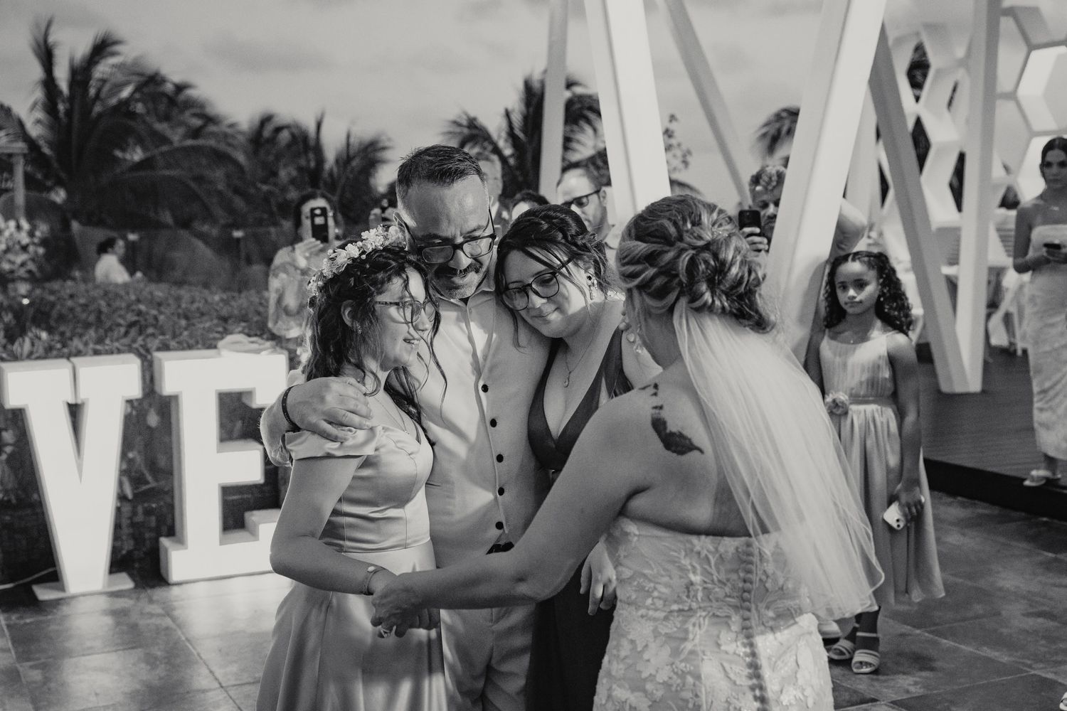 Black and white photo of bride and groom embracing children near LOVE sign at reception.