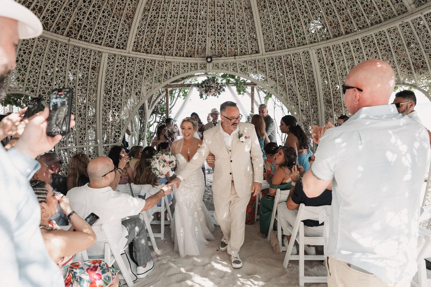 Newlywed couple walks down the aisle through cheering guests inside an ornate domed wedding venue.