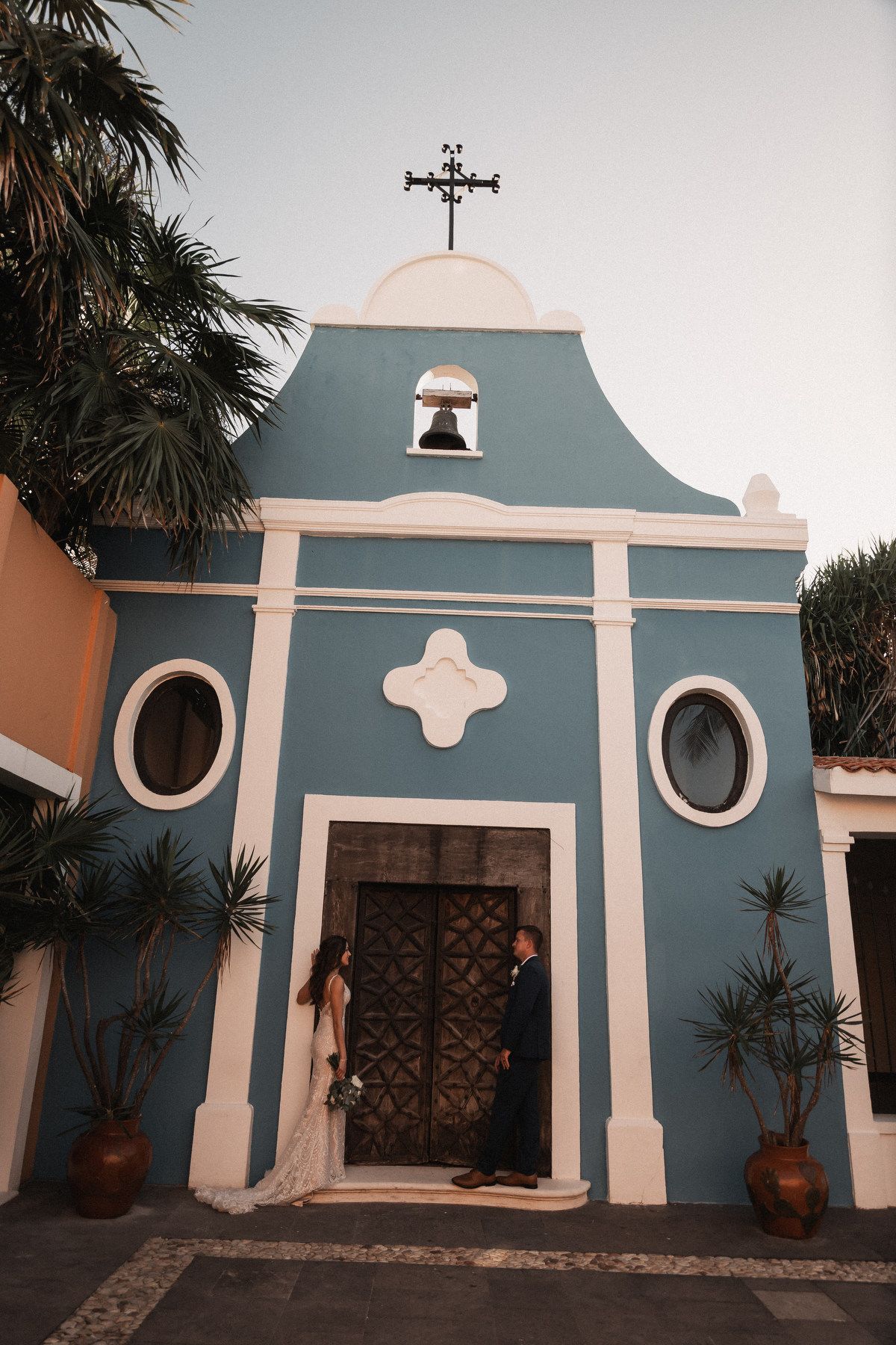 Bride and groom share intimate moment at entrance of charming blue and white chapel at dusk destination wedding.