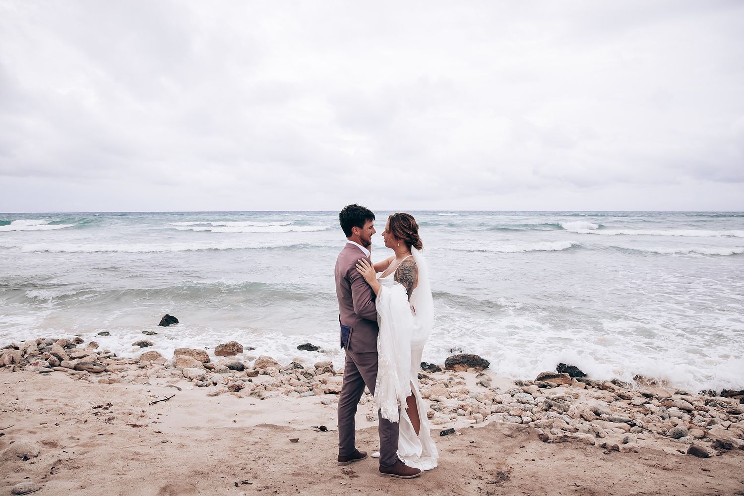 Couple walks along rocky oceanfront beach in wedding attire.
