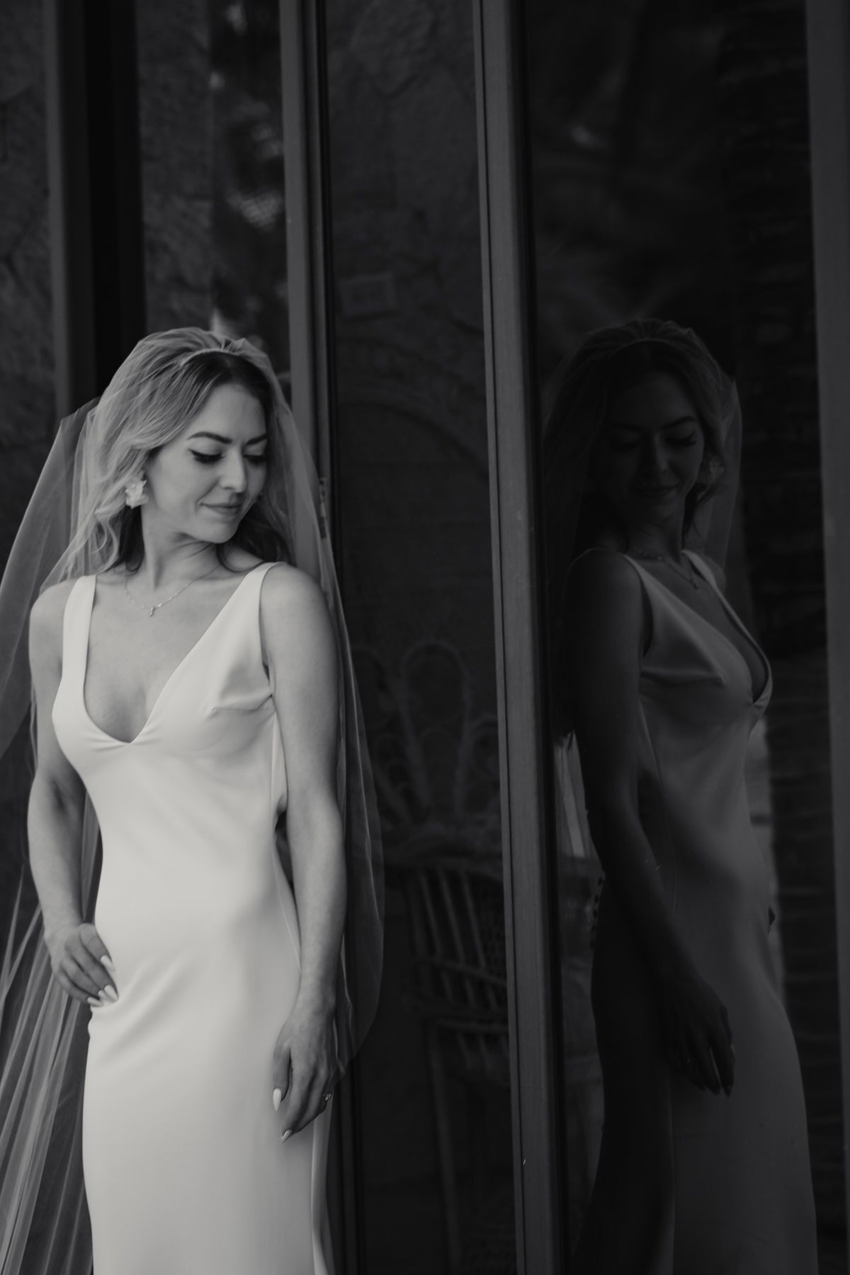 Black and white portrait of a blonde bride in a sleek white gown and veil leaning against a glass wall.