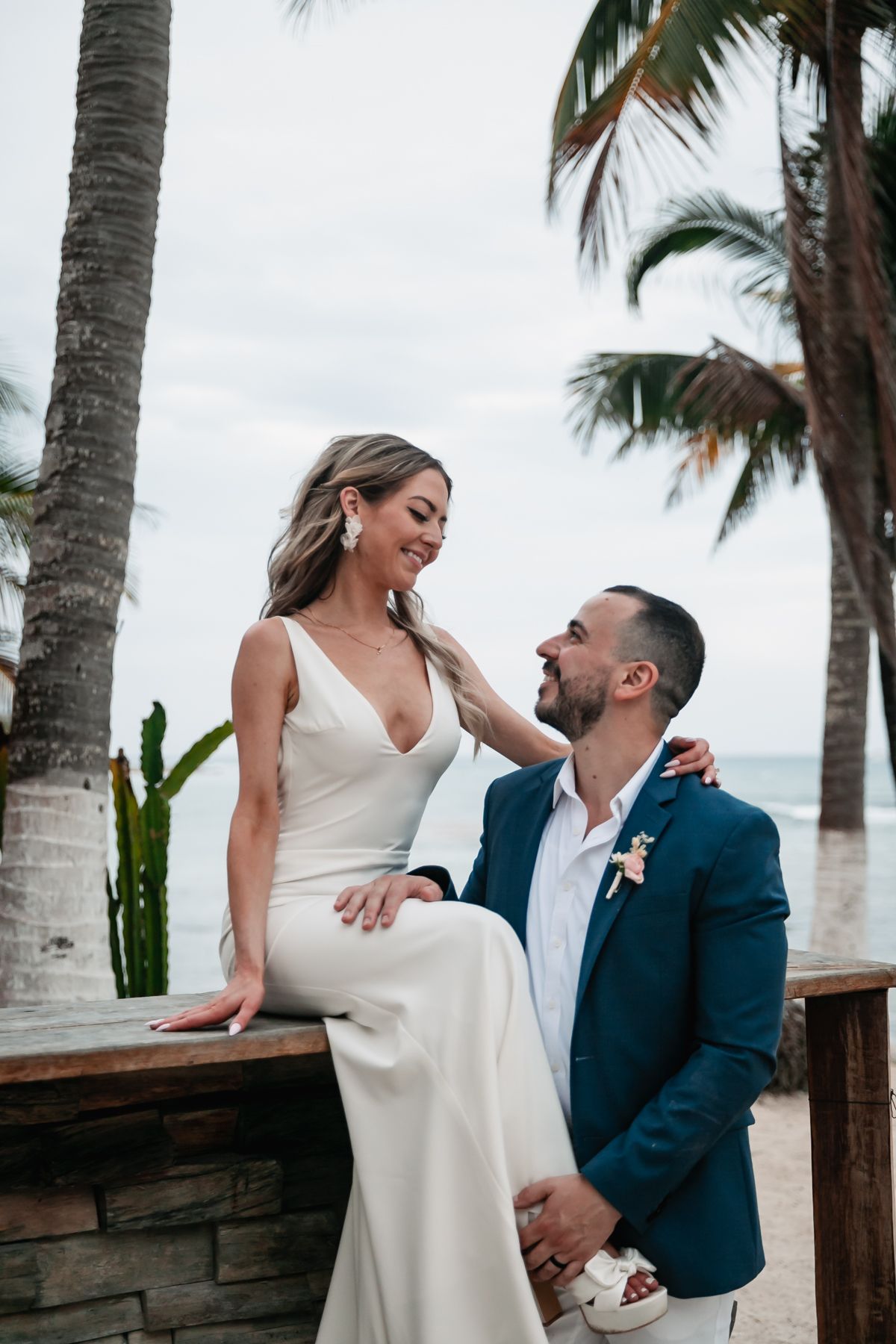 Bride in white gown and groom in blue suit share a romantic moment by palm trees on a beach.