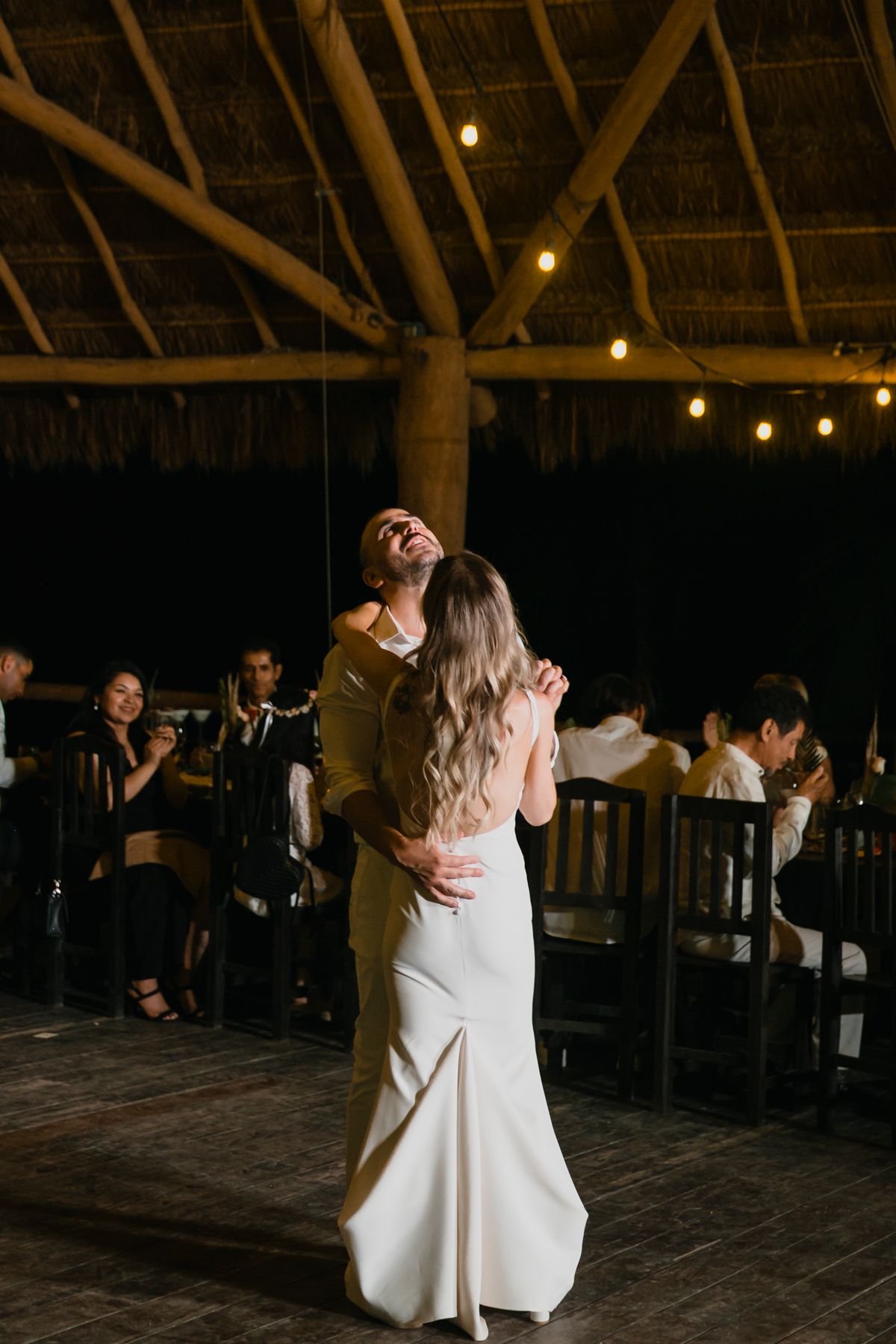 Groom laughs joyfully while dancing with his bride under string lights at a tropical wedding reception.