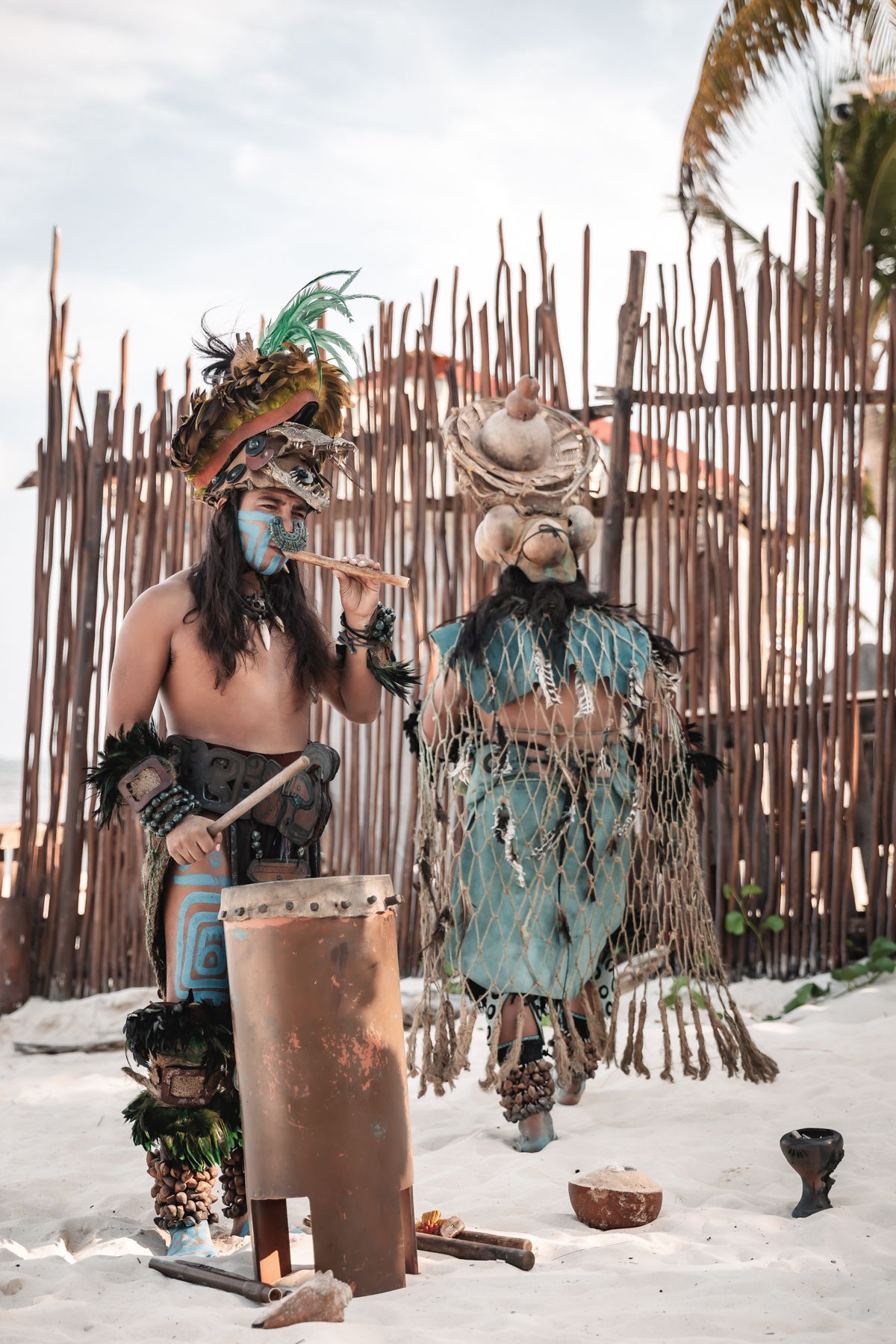 Two performers in traditional Mayan costumes with face paint and headdresses playing instruments on a sandy beach.