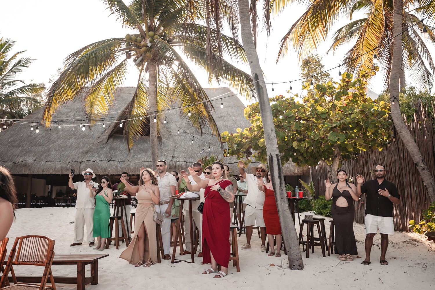 Guests celebrate at a tropical beach wedding reception with palm trees and a thatched-roof venue at sunset.