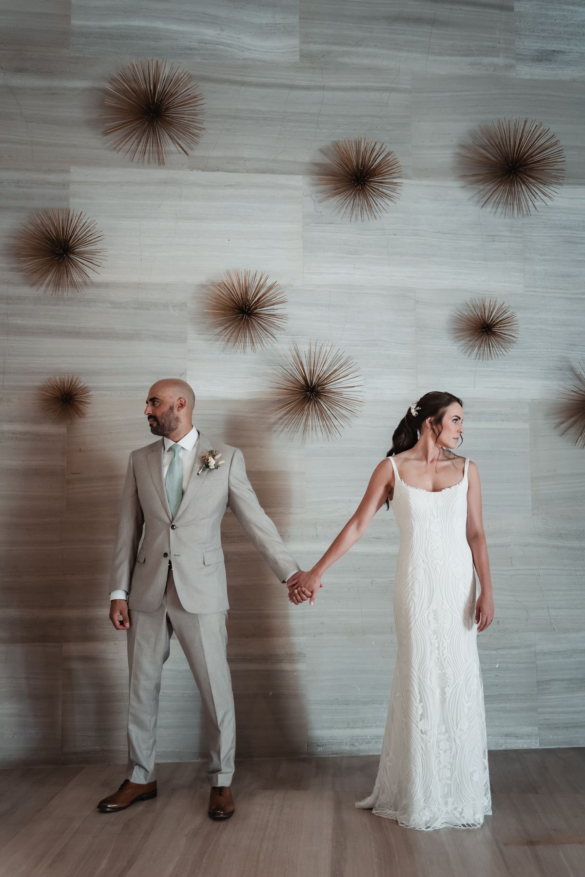 Bride in white lace gown and groom in tan suit hold hands before a modern wall with gold starburst decor.