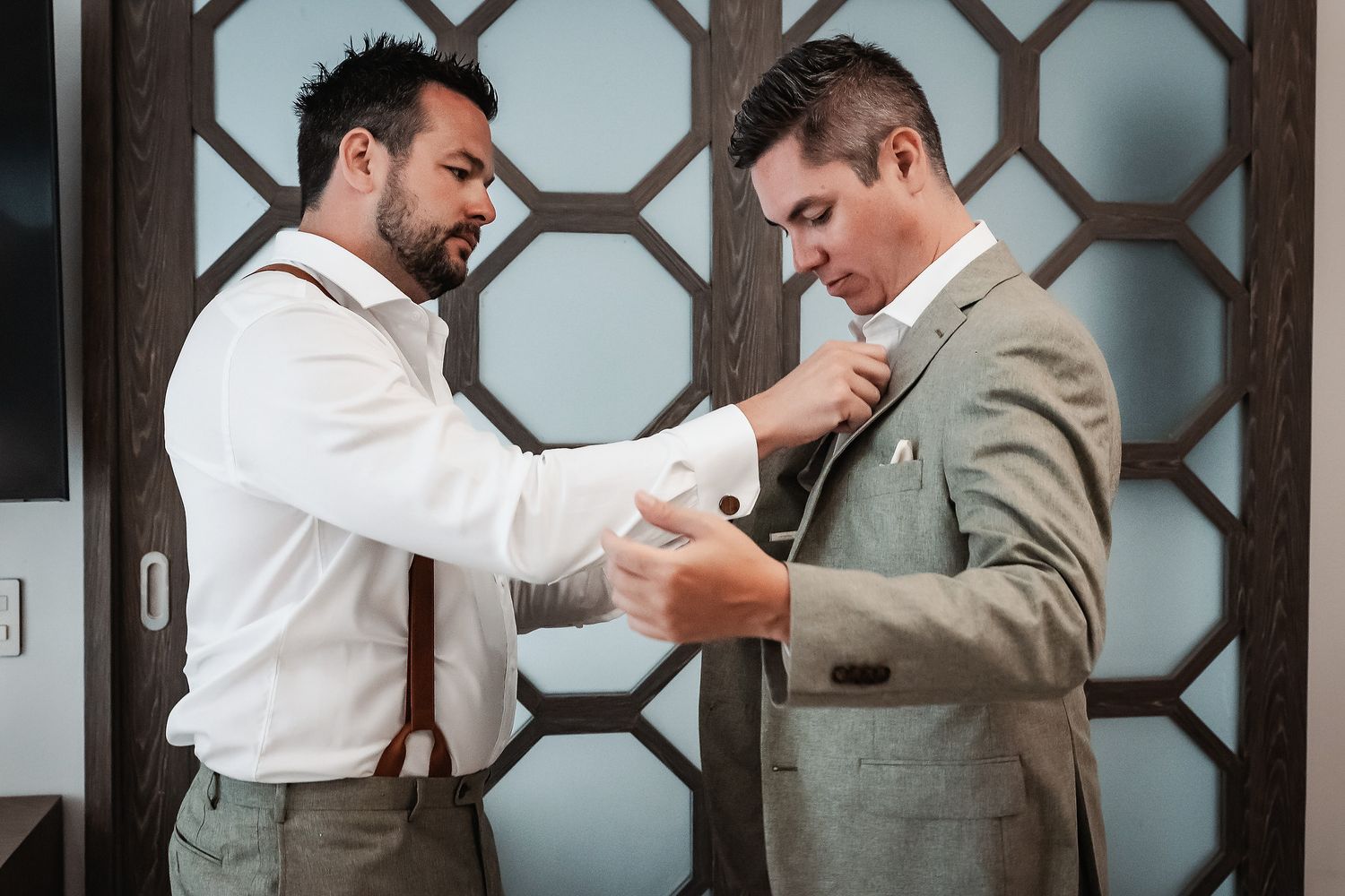 Two groomsmen helping each other get dressed, one adjusting a tie while the other fixes a cufflink before a wedding.