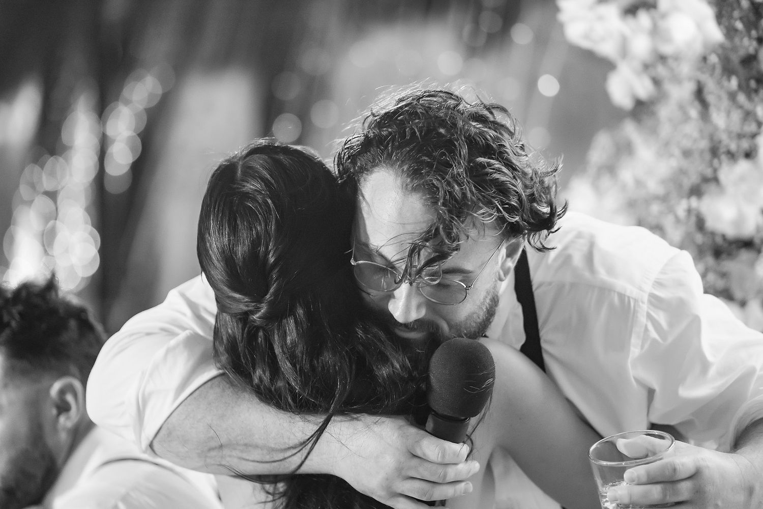 Black and white photo of a man with curly hair and glasses sharing an emotional hug at an outdoor wedding.