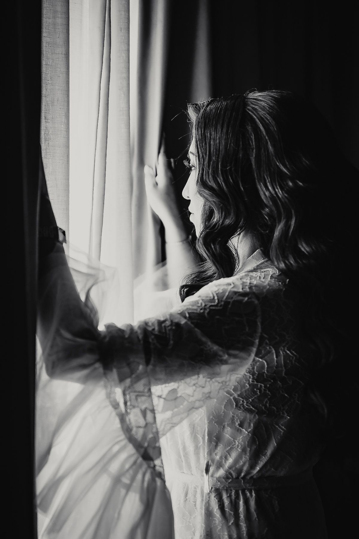 Black and white photo of a woman in lace robe gazing out a window with sheer curtains.