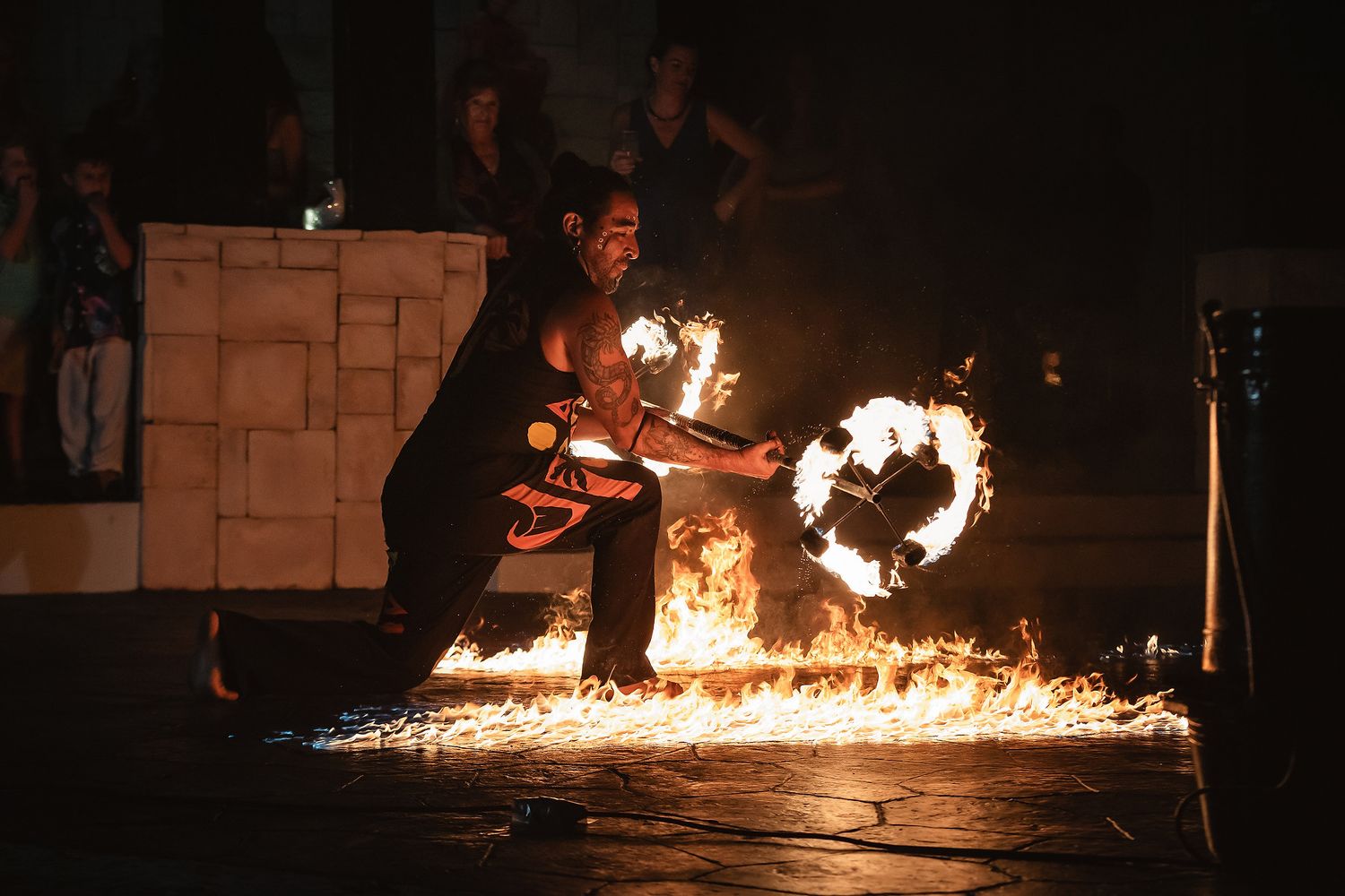 Fire performer kneeling on stage manipulating flaming rings during a dramatic nighttime fire dance show.