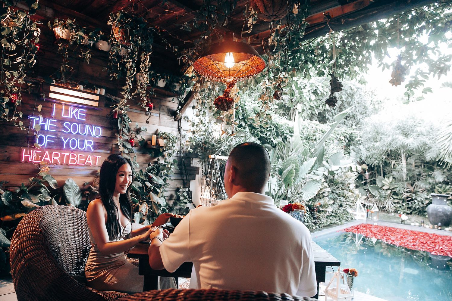 Romantic couple dining by a pool under lush greenery with a neon sign reading I Like The Sound Of Your Heartbeat.