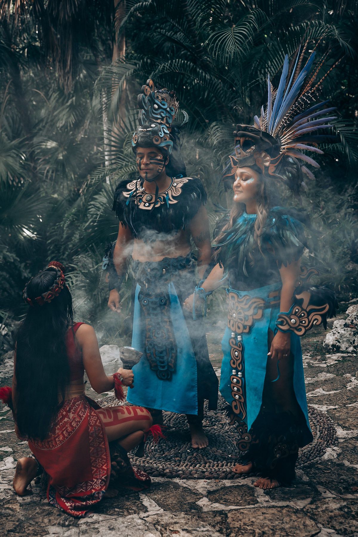 Three people in elaborate Mayan ceremonial costumes with feathered headdresses perform a ritual with smoke in a jungle setting.
