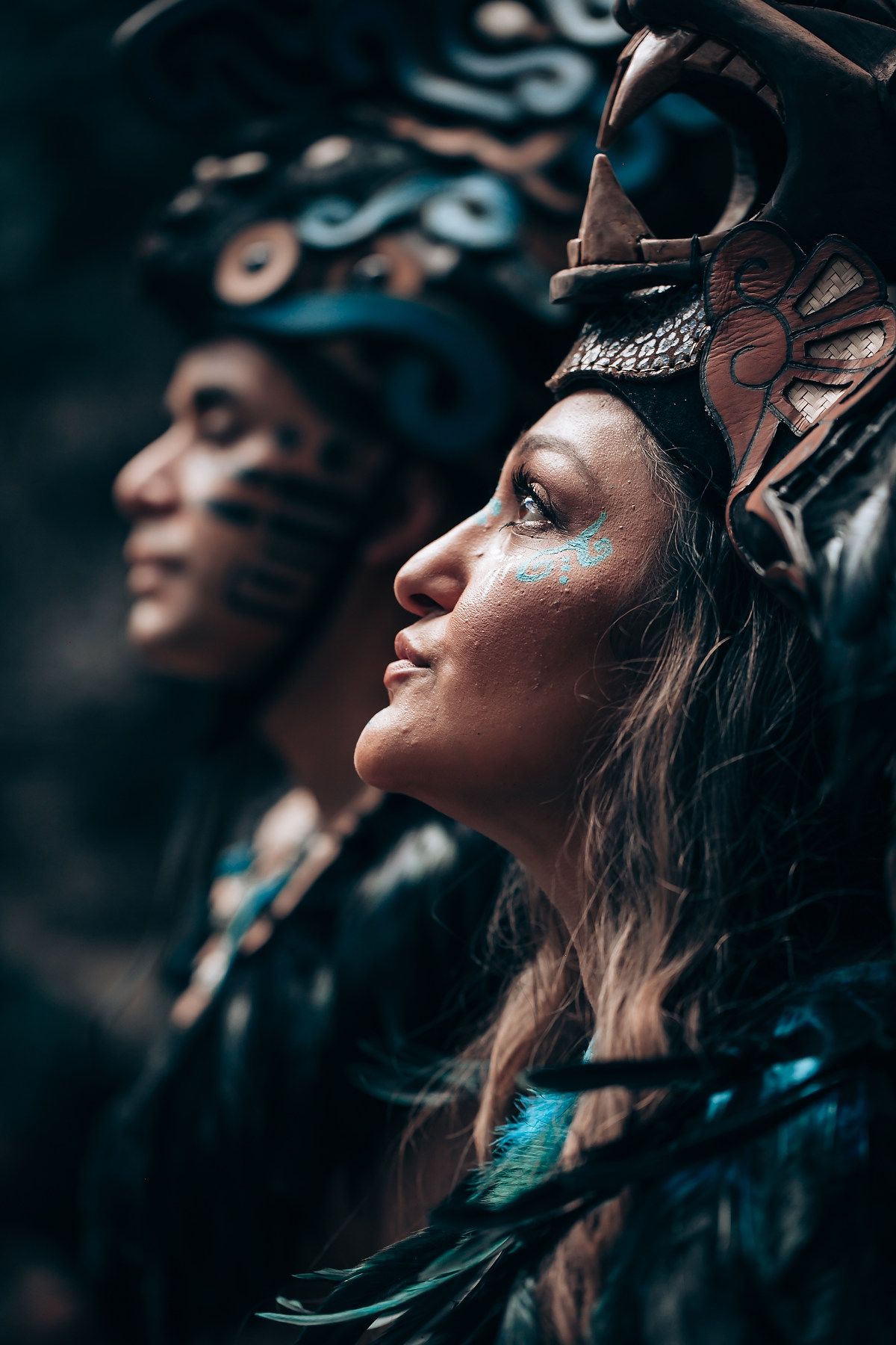 Two women in profile wearing elaborate Mayan headdresses and turquoise face paint in a dramatic forest scene.