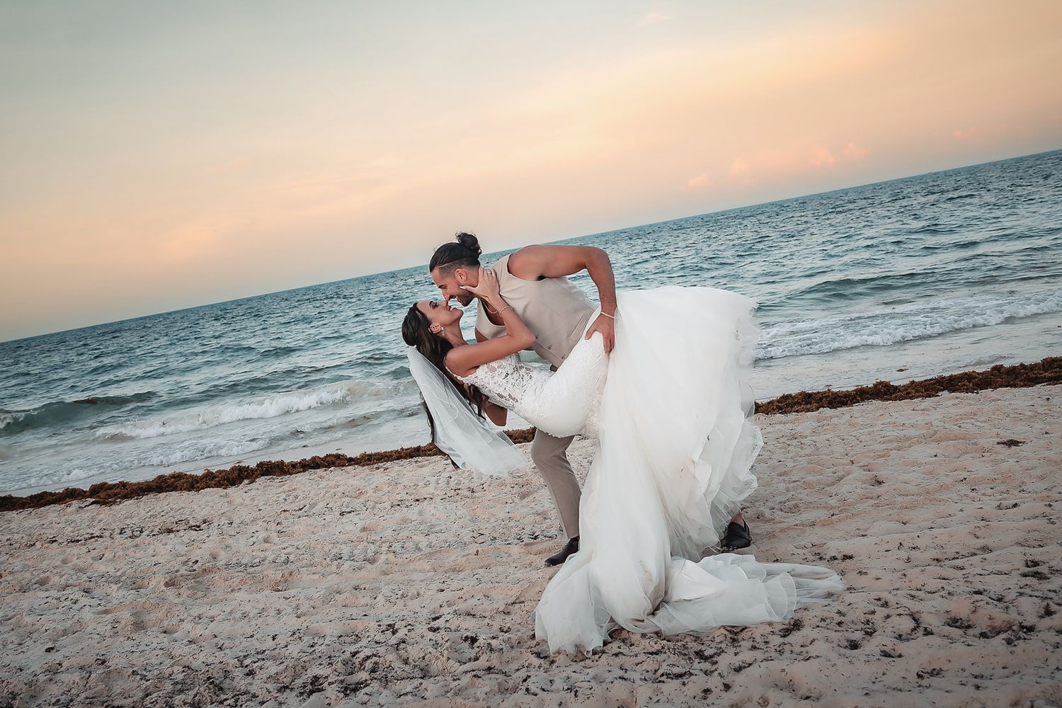 Bride and groom share a romantic dip kiss on a sandy beach at sunset with ocean waves in the background.