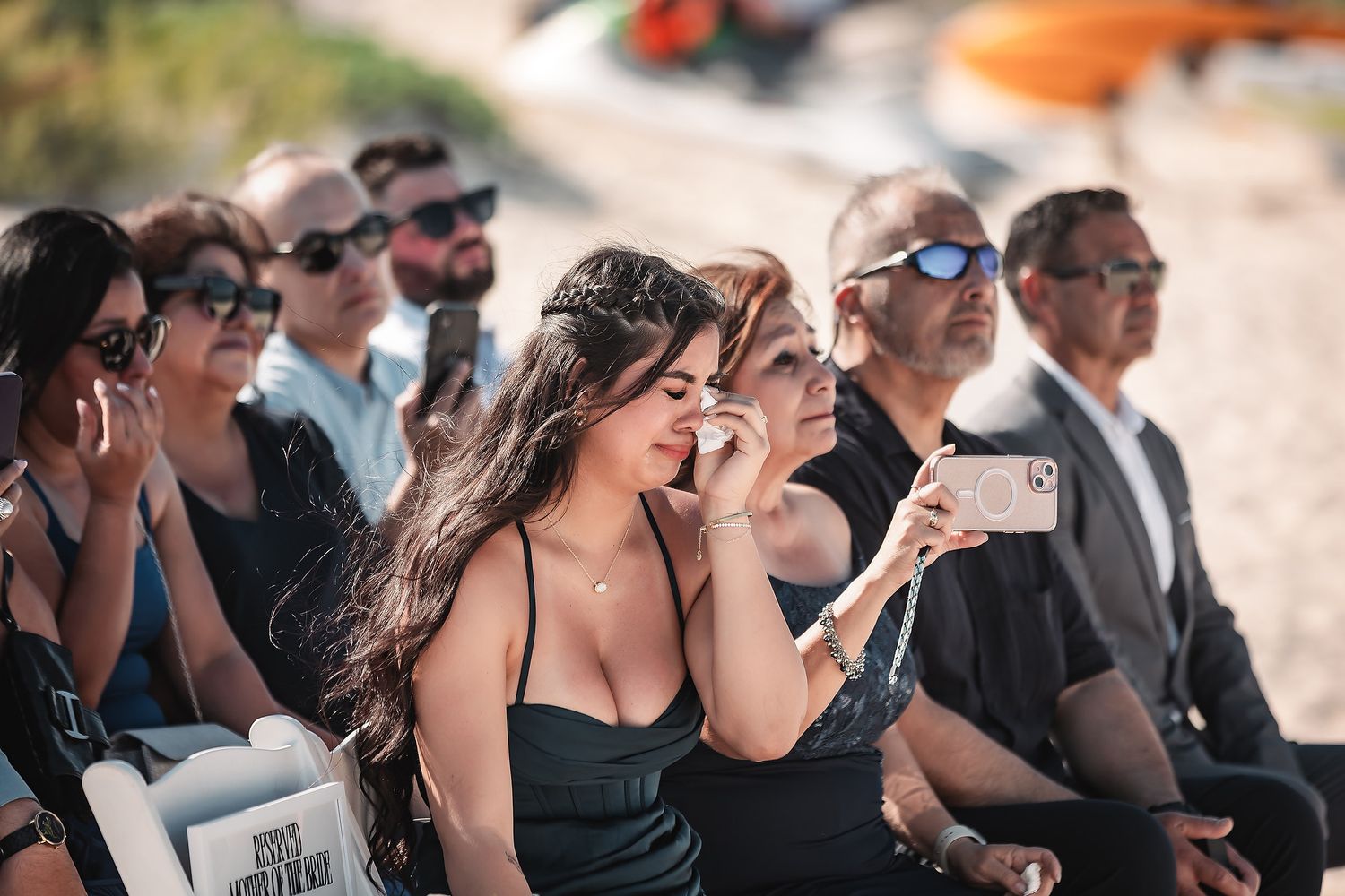 Emotional woman in black dress crying at outdoor wedding ceremony while guests sit in background.