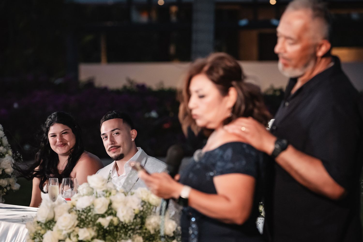 Elegant wedding reception with two couples, featuring floral arrangements and formal attire at an upscale evening event.