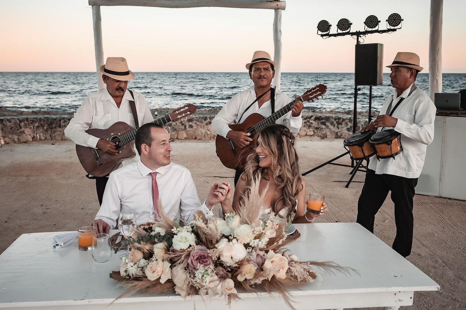 Bride and groom sit at decorated table on beach while mariachi band performs at sunset wedding reception.