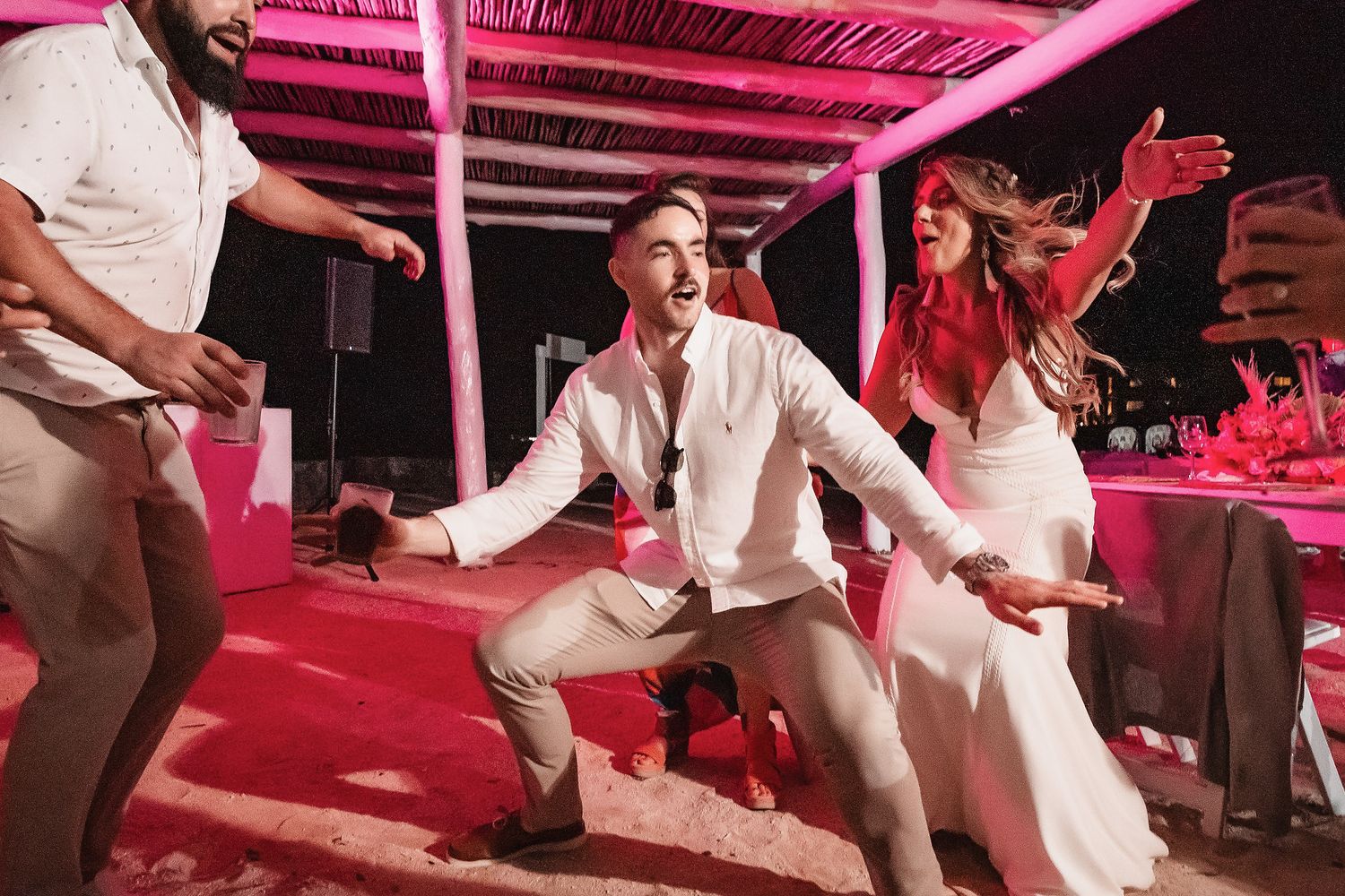 People dancing joyfully at an outdoor wedding reception under pink lights at night.