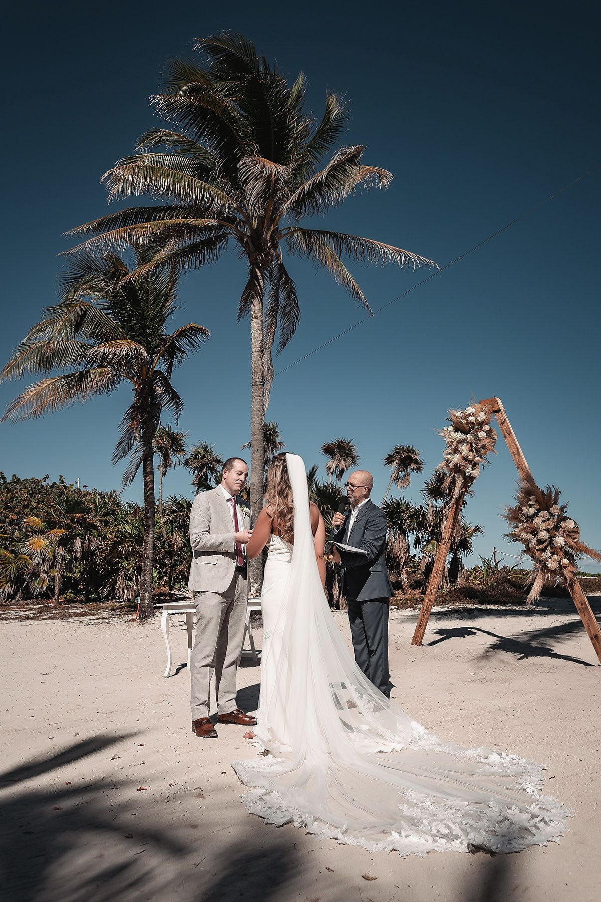 Bride and groom exchange vows at a tropical beach wedding ceremony with palm trees and a wooden arch.