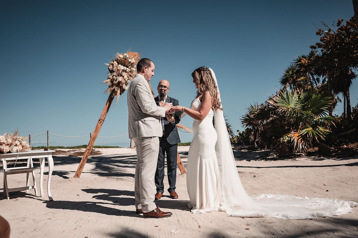 Couple exchanging vows on a sandy beach with a boho floral arch, officiant, and palm trees under a clear blue sky.