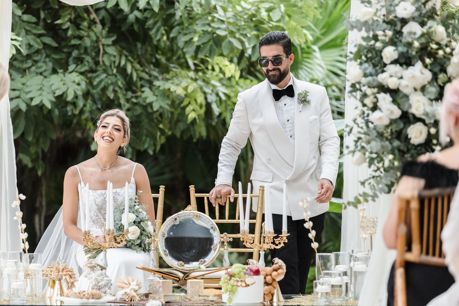 Bride and groom smiling at elegant outdoor wedding reception with white floral decor and gold accents.