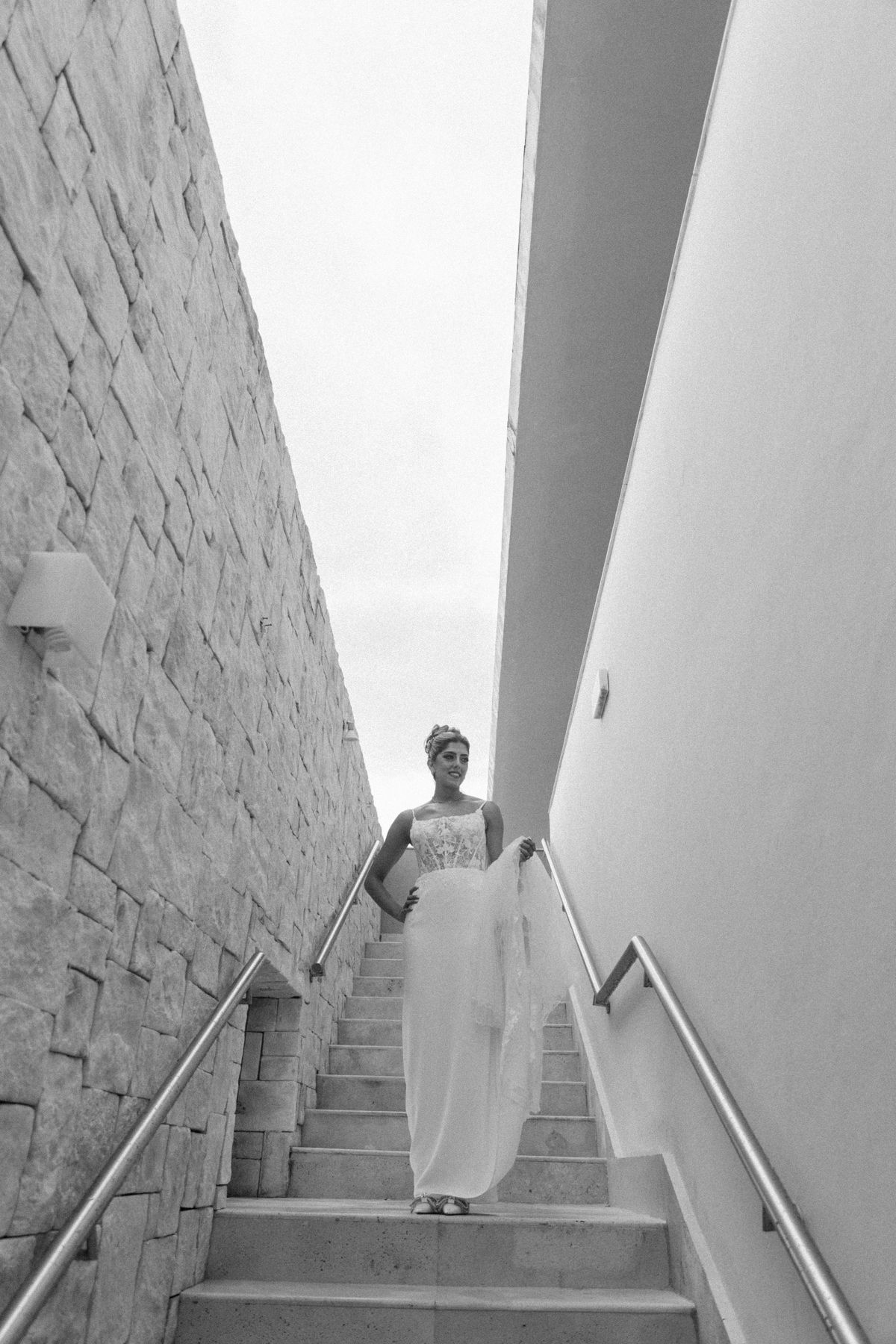 Bride in white gown descending stone staircase between walls, black and white wedding photography.