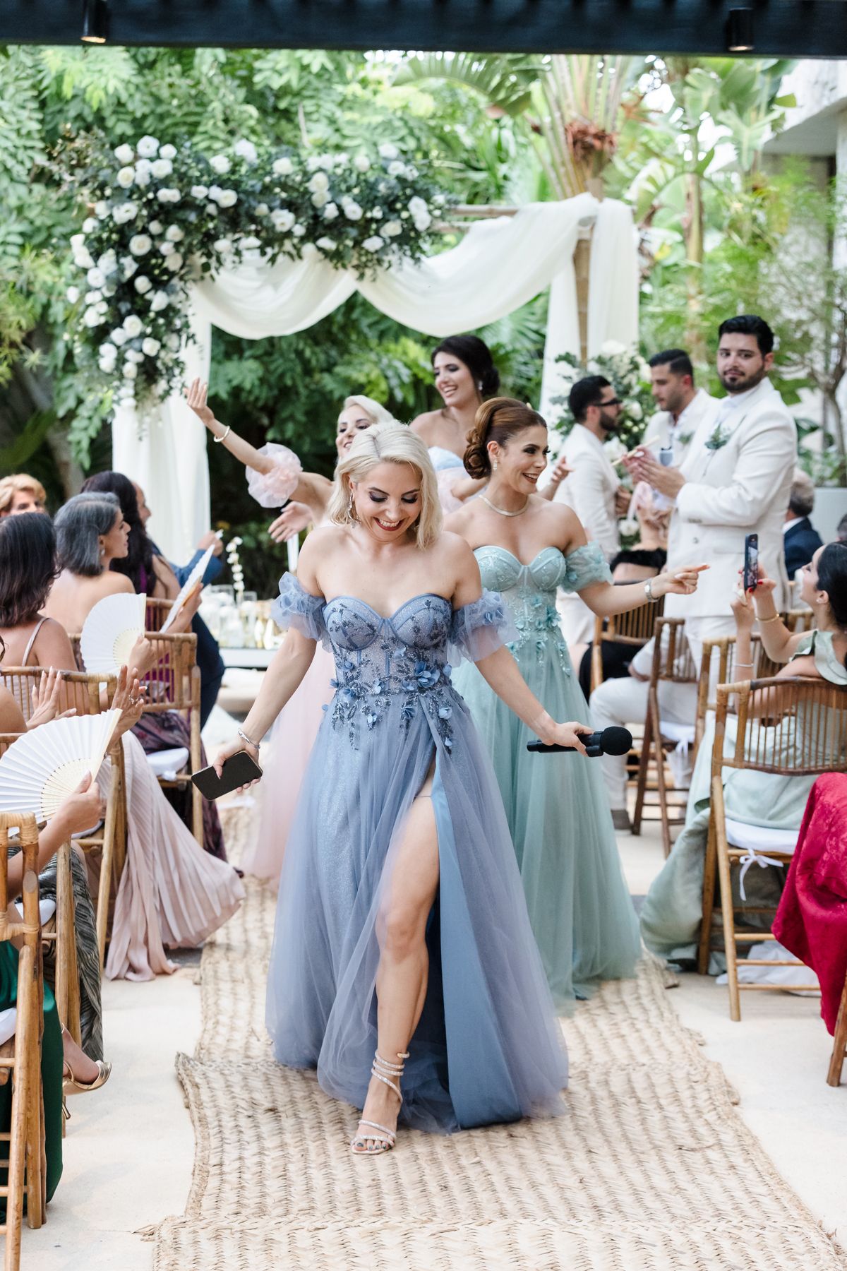 Bridesmaids in blue off-shoulder tulle gowns walk down an outdoor wedding aisle surrounded by guests.