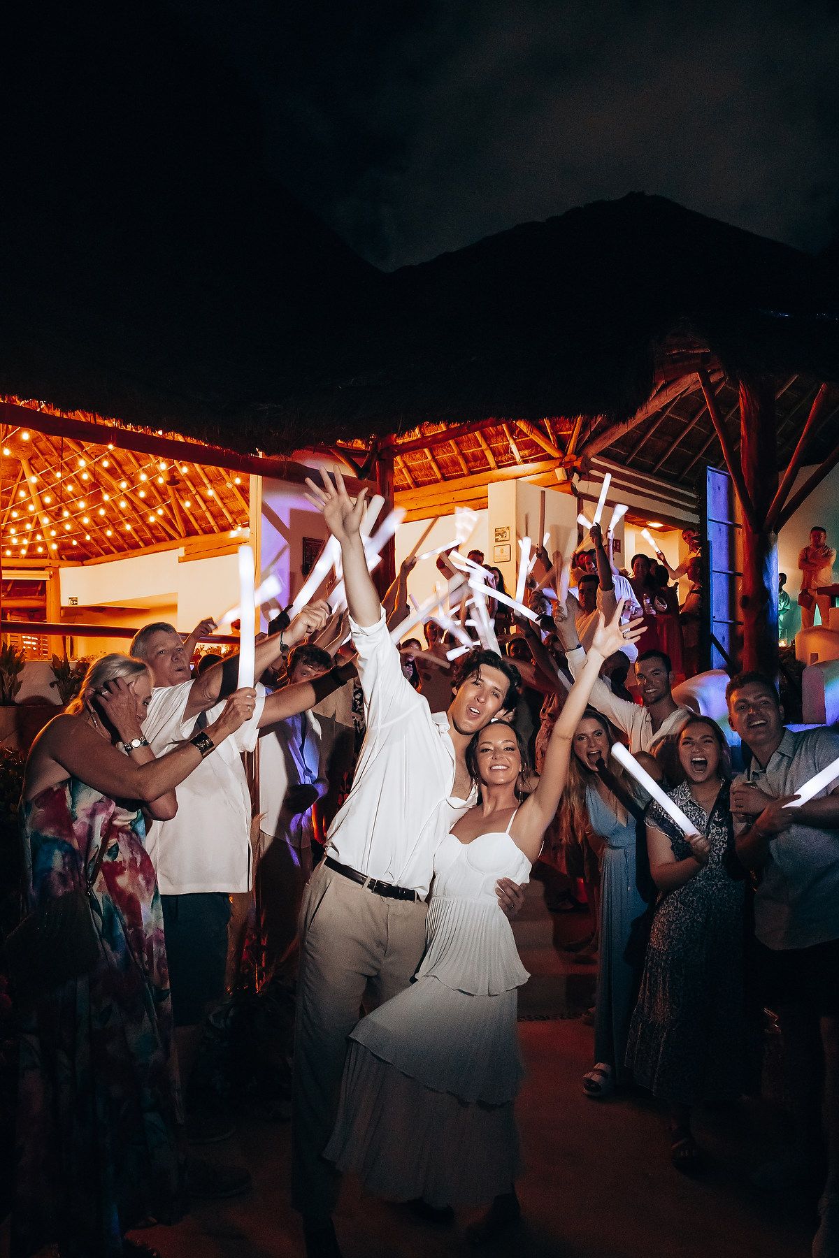 Wedding party celebrates on dance floor with glow sticks under string lights.