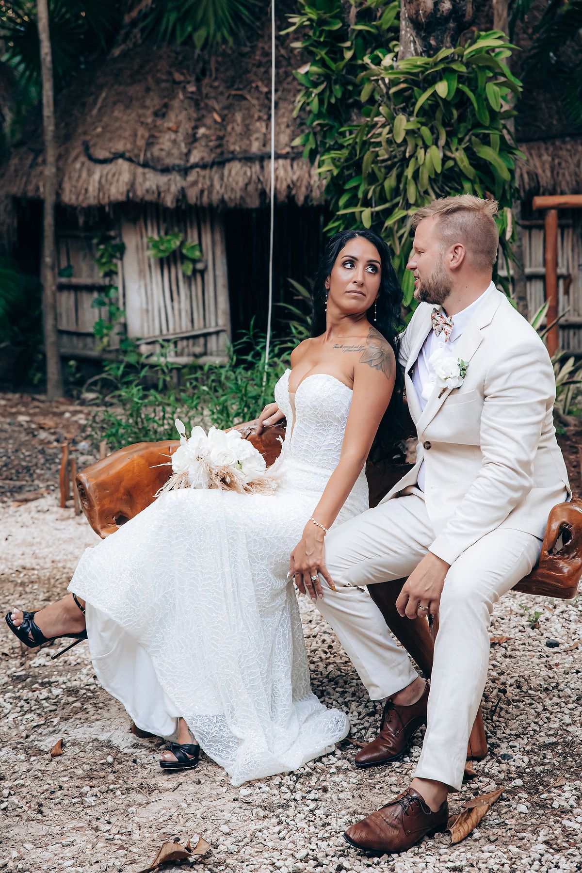 Newlyweds in white attire sitting together on rustic wooden furniture in a garden setting.