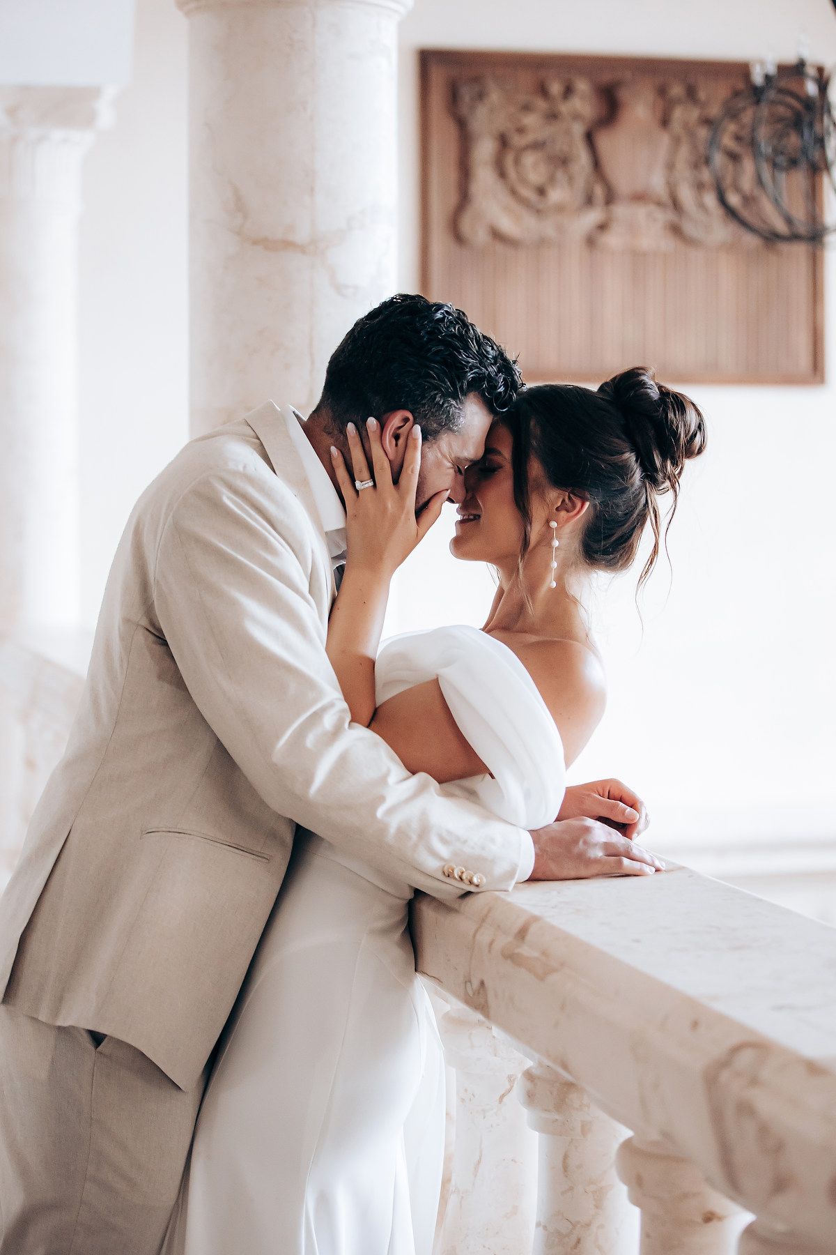 A couple in white attire share an intimate moment in an elegant venue with white columns.
