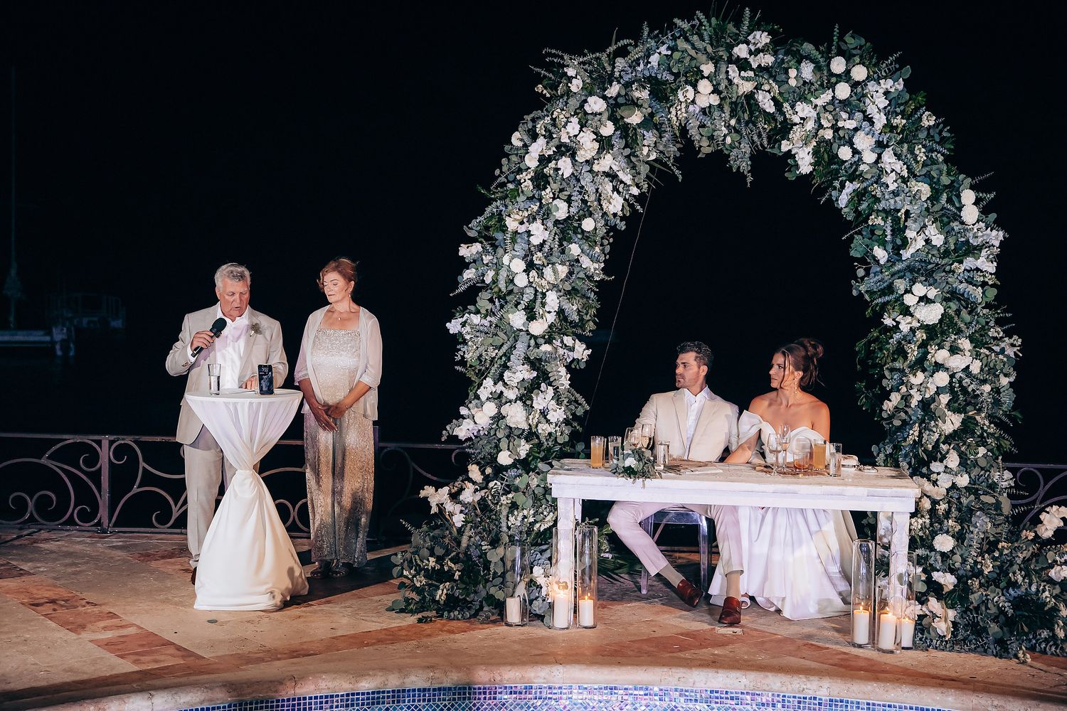 A romantic outdoor wedding ceremony setup at night with a floral arch and white decorations.