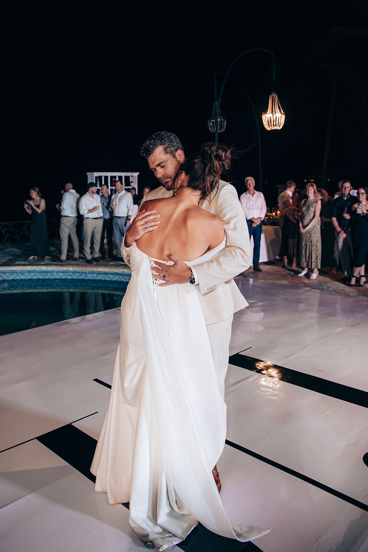A couple shares an intimate first dance moment at their wedding reception on a white dance floor with guests watching.