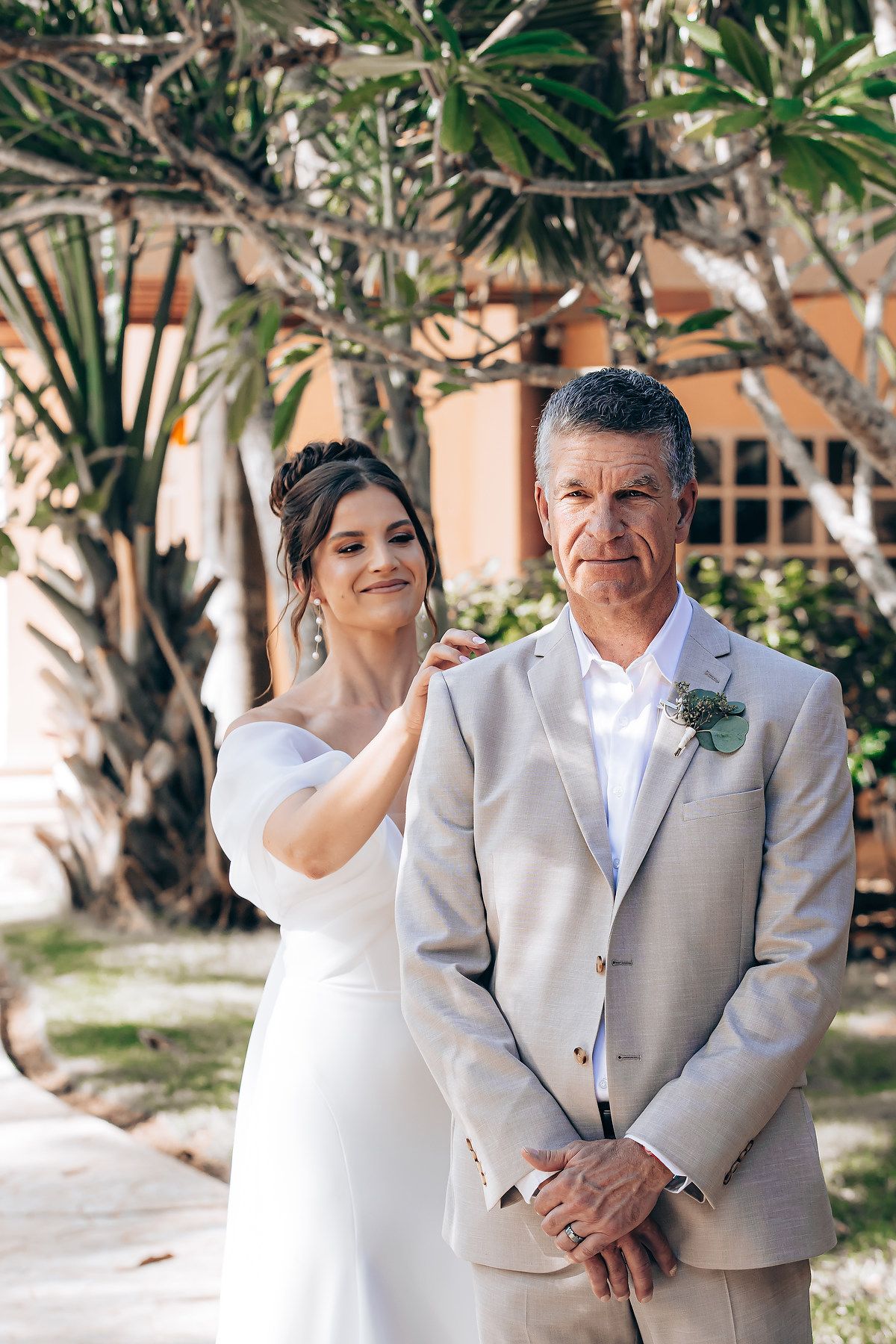 A couple in wedding attire share a tender moment in an outdoor garden setting with tropical foliage in the background.