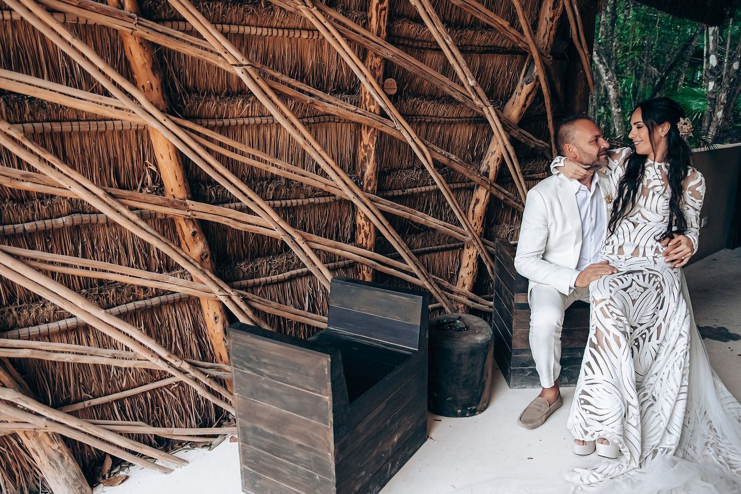 A couple in elegant attire poses together near a modern architectural structure with woven bamboo details.