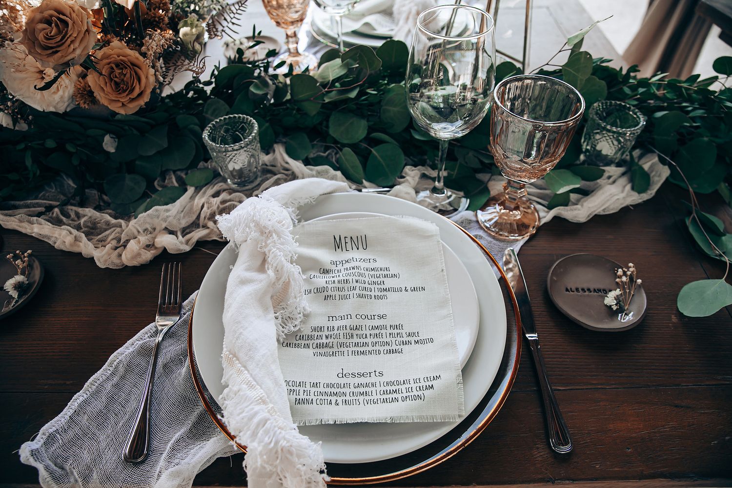 Elegant wedding table setting with menu card on white plate, eucalyptus greenery and wine glasses on dark wooden table.