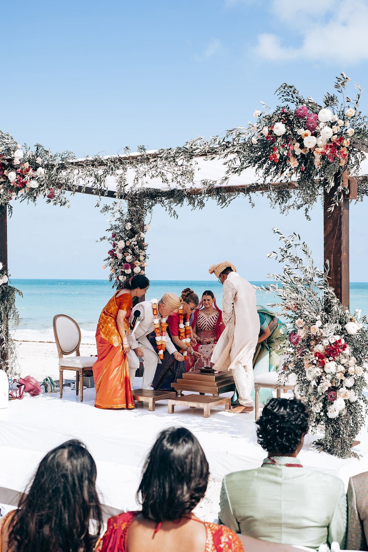 Intimate beach wedding ceremony with guests watching under a flower-adorned wooden pergola.