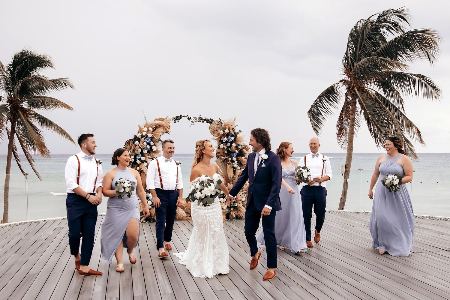 Panoramic view of a beach wedding party posing on a wooden boardwalk with palm trees in the background.