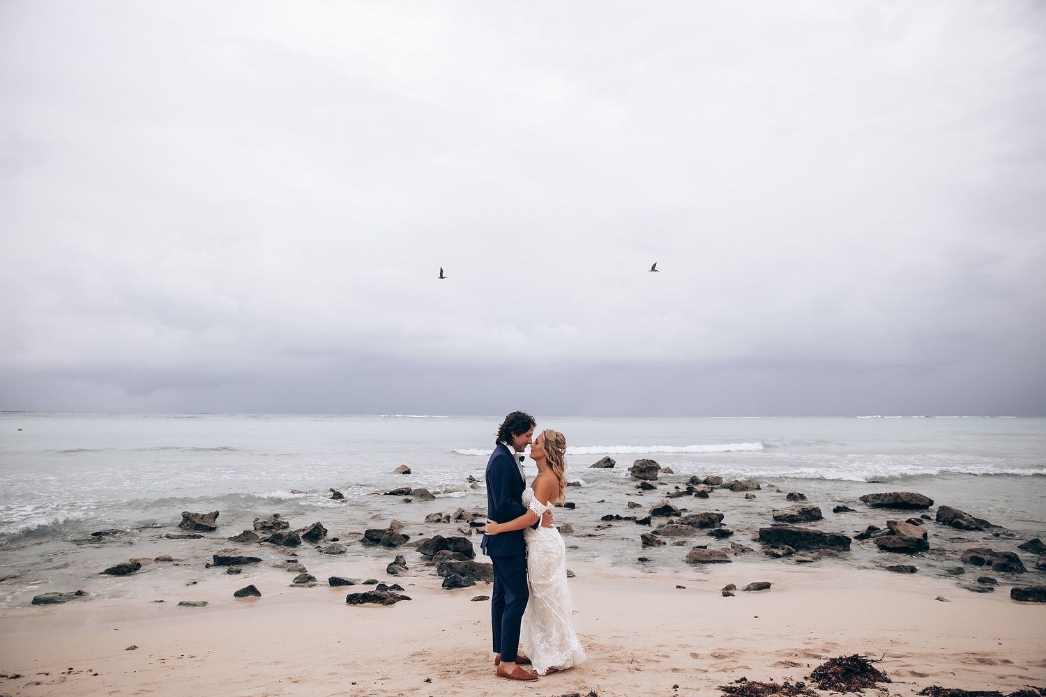 A couple embraces on a rocky beach shore during an overcast day.