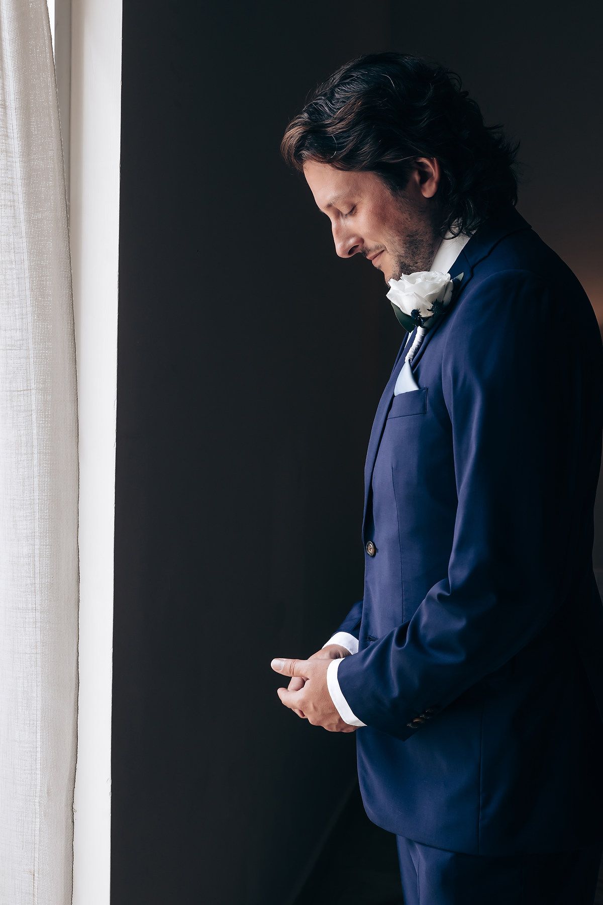 A person in a navy suit checks their phone while standing near a window.