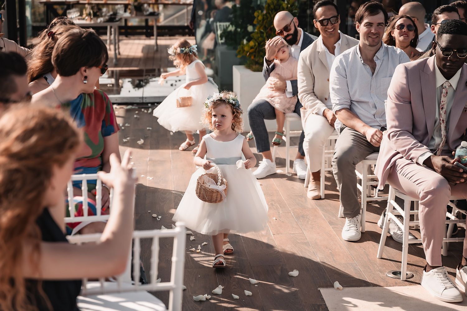 Wedding ceremony guests seated in white chairs at an outdoor venue with natural lighting.