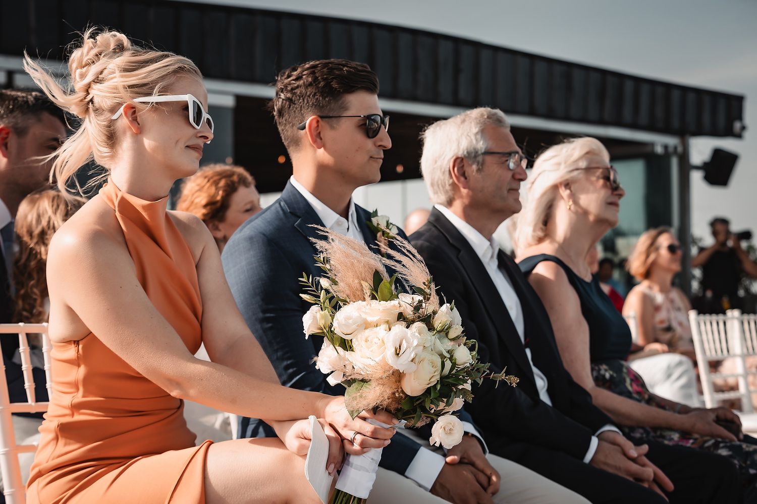 Wedding guests seated in white chairs watching outdoor ceremony.