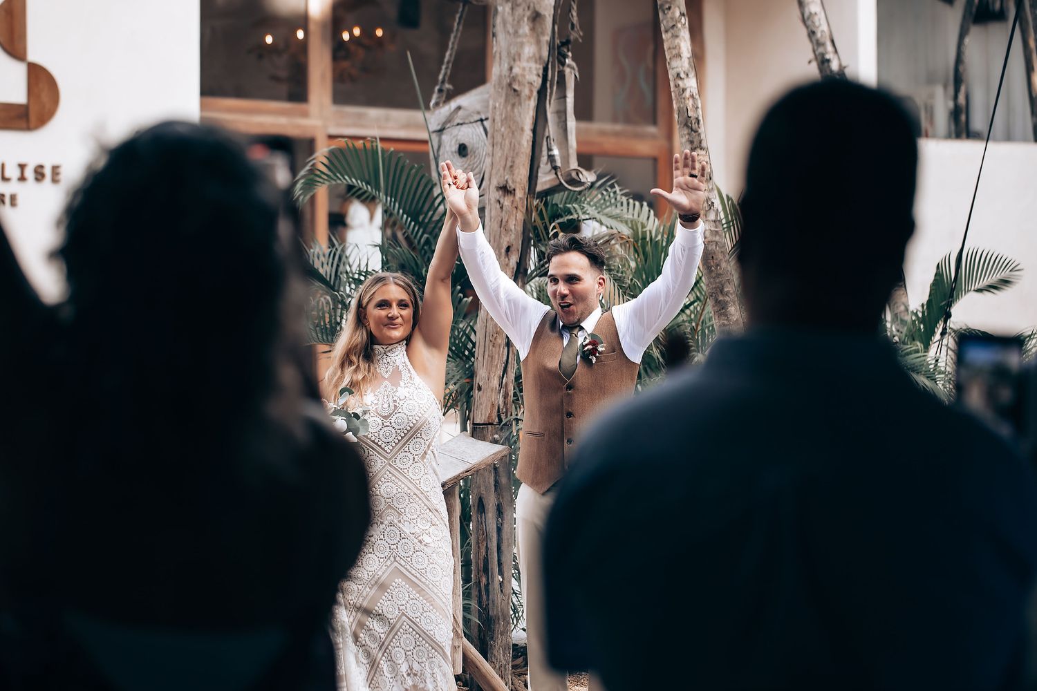 A bride in a white lace wedding dress dances joyfully with raised arms at an outdoor reception celebration.