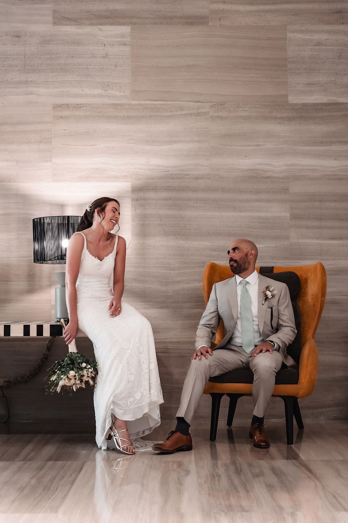 A couple shares a joyful moment in a modern setting with grey walls, where she sits on a bench and he in an orange chair.