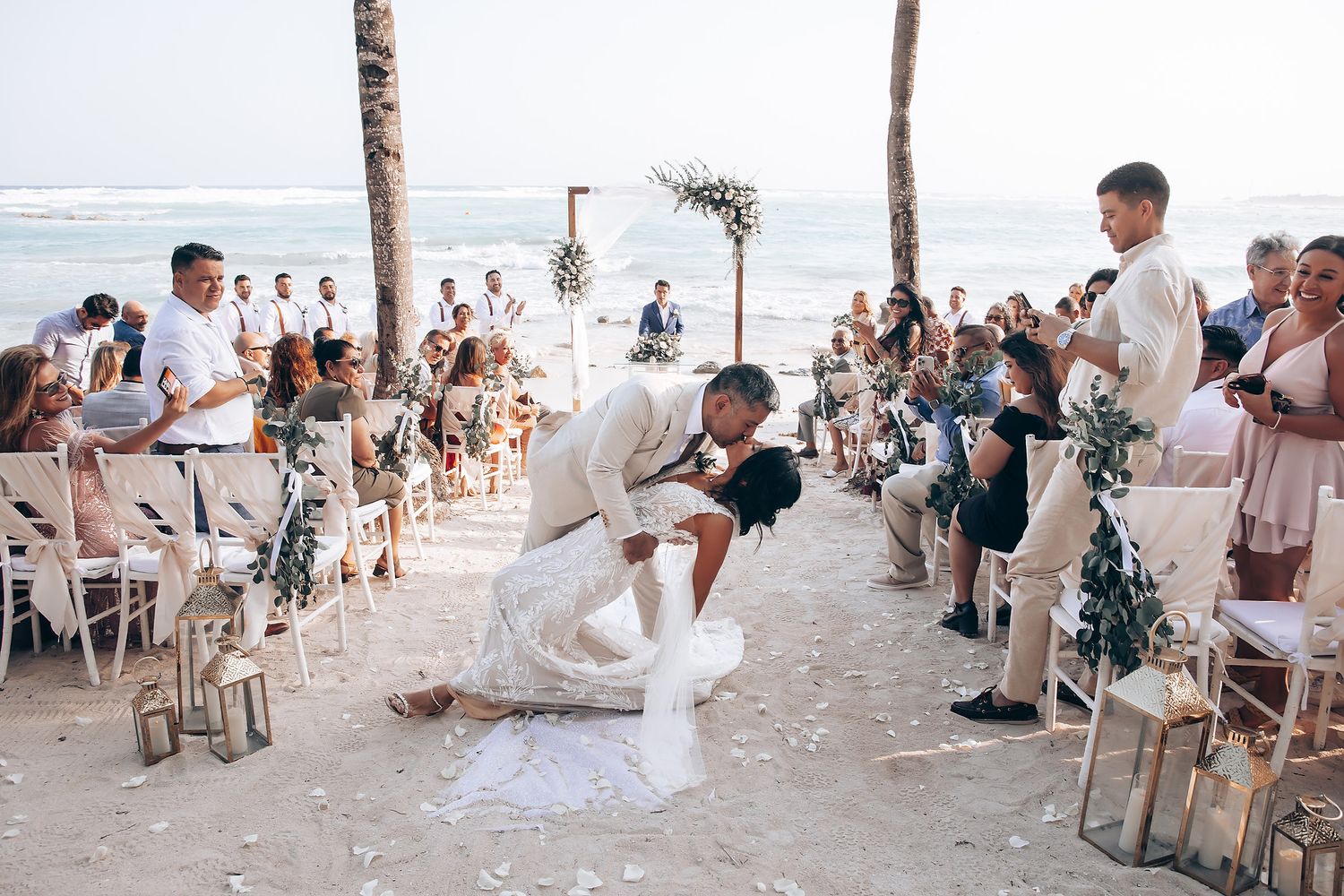 Wedding guests gather on a sunny beach with palm trees and white chairs for an oceanside ceremony.